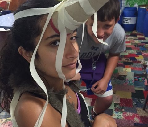 A girl with a paper towel roll taped to her head, a smiling boy is hiding behind her.