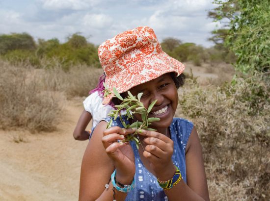 A girl wearing hat in center of photo holding leaf up to her eye.