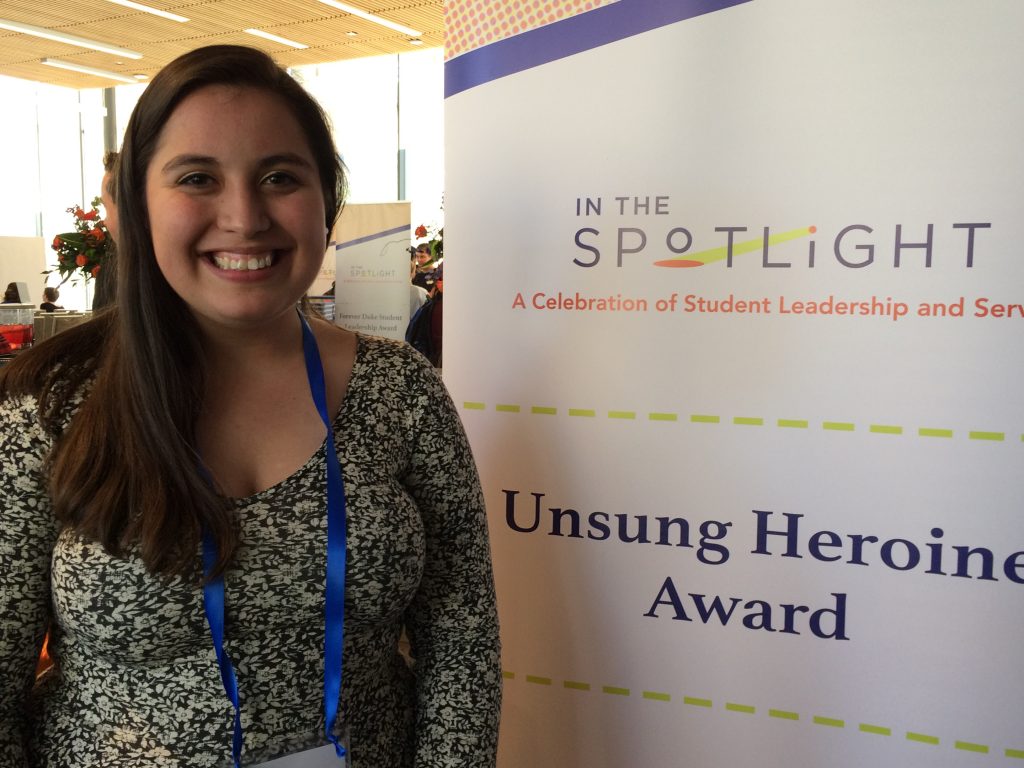 young woman in a dress poses beside banner with text that reads Unsung Heroine Award