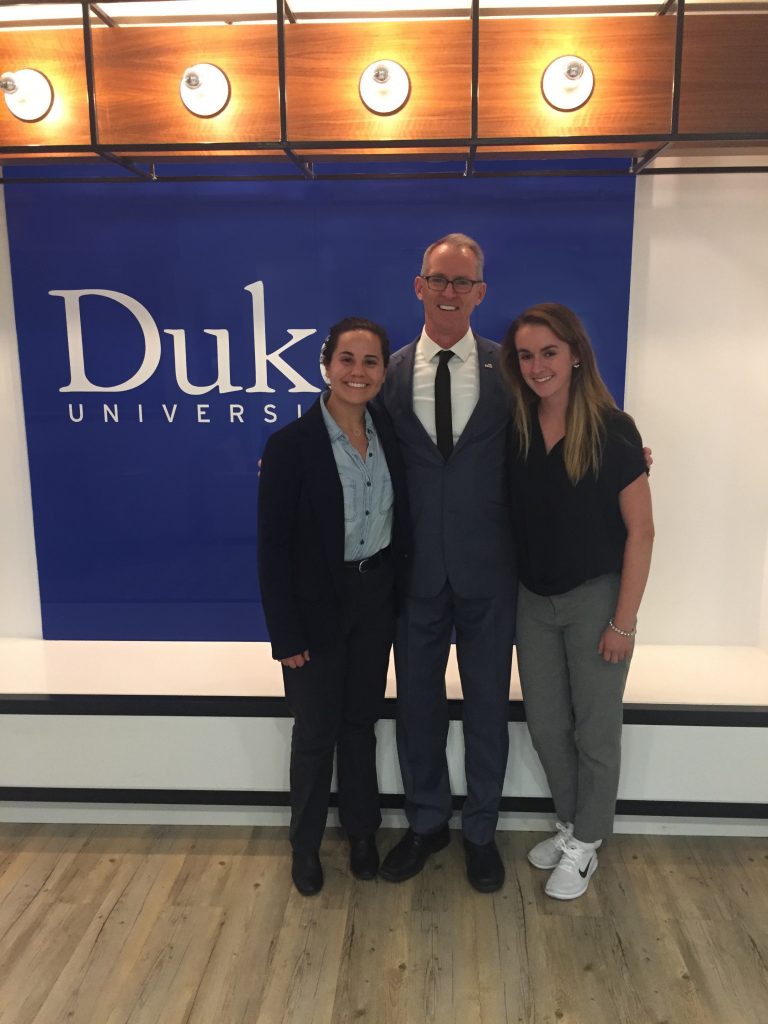 three poeple posing for camera in front of large blue banner with the word Duke on it