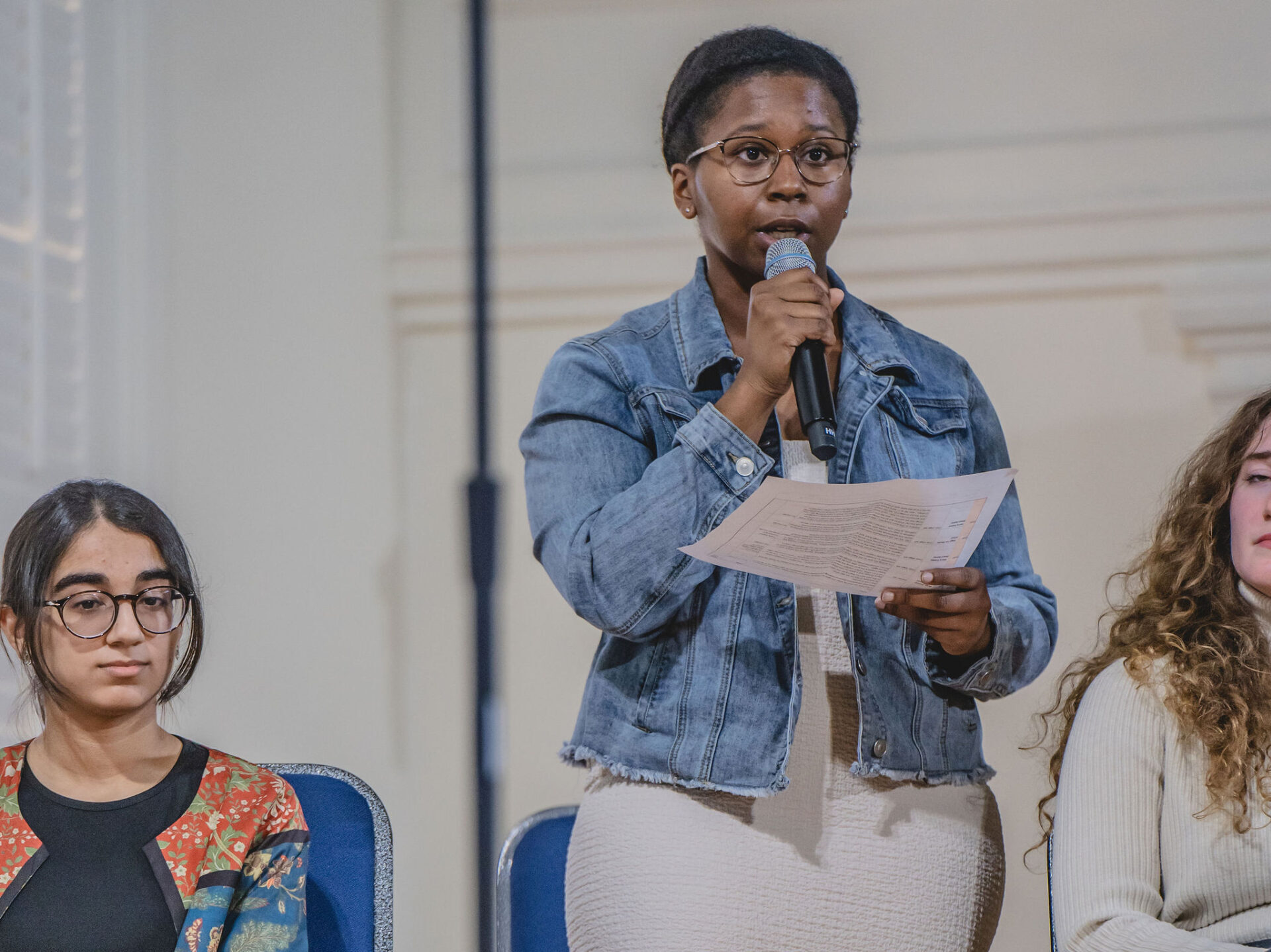 A young woman in a denim jacket reads from a piece of paper