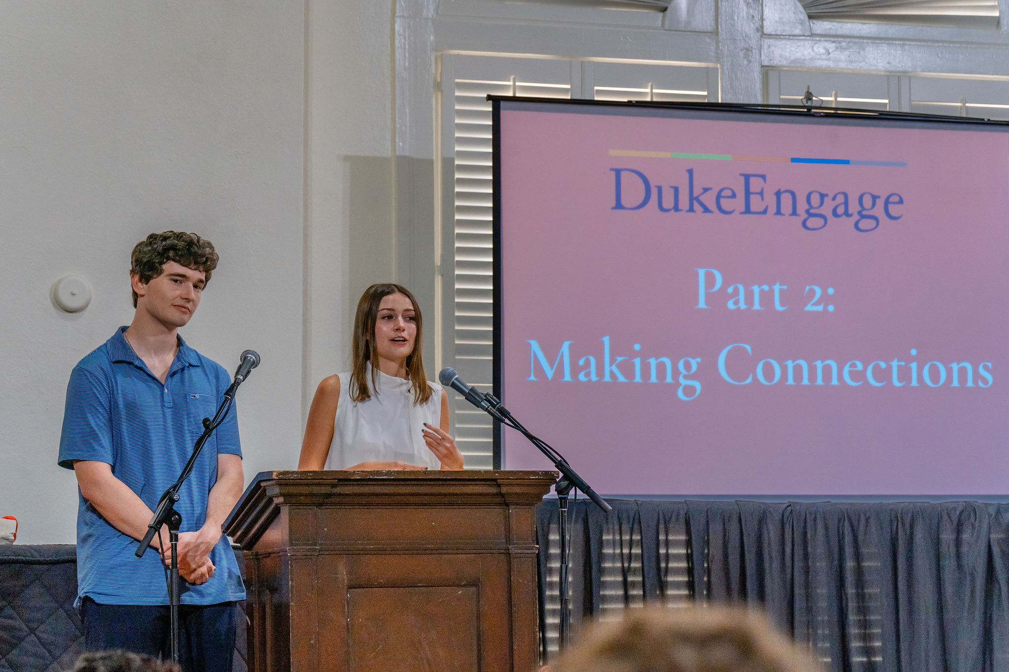 Two students stand at a podium with a slide in the background reading DukeEngage Part 2: Making Connections