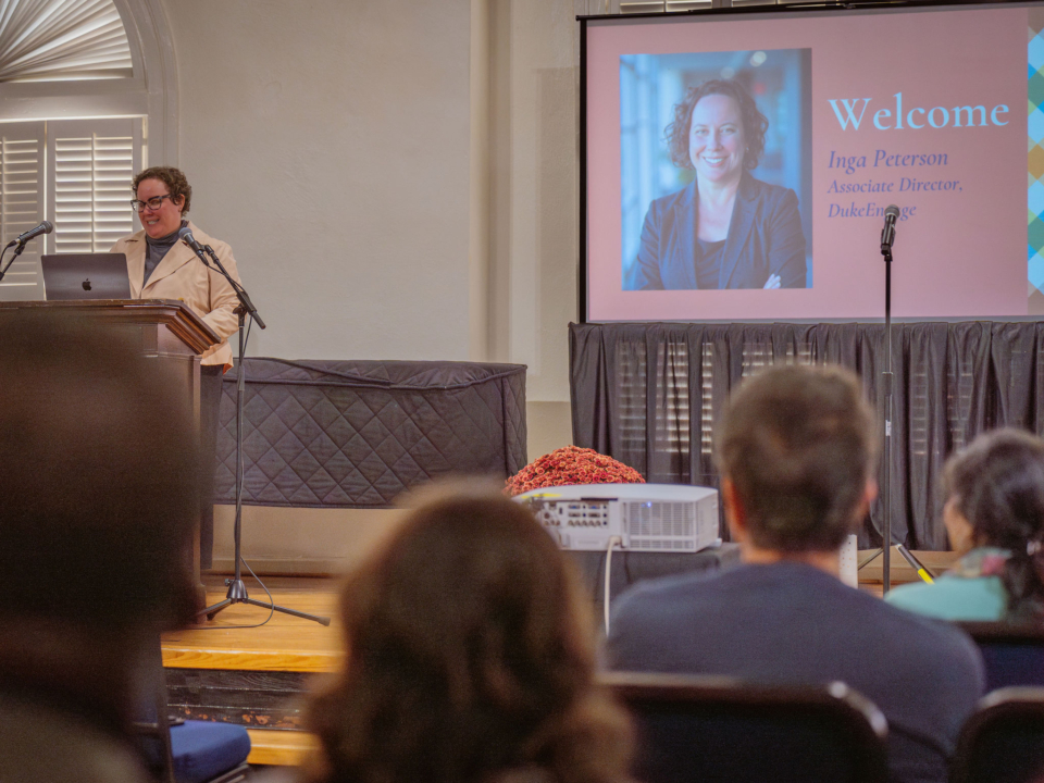 A woman stands at a podium, addressing an audience, smiling. A slide reads "Inga Peterson, Associate Director, DukeEngage"